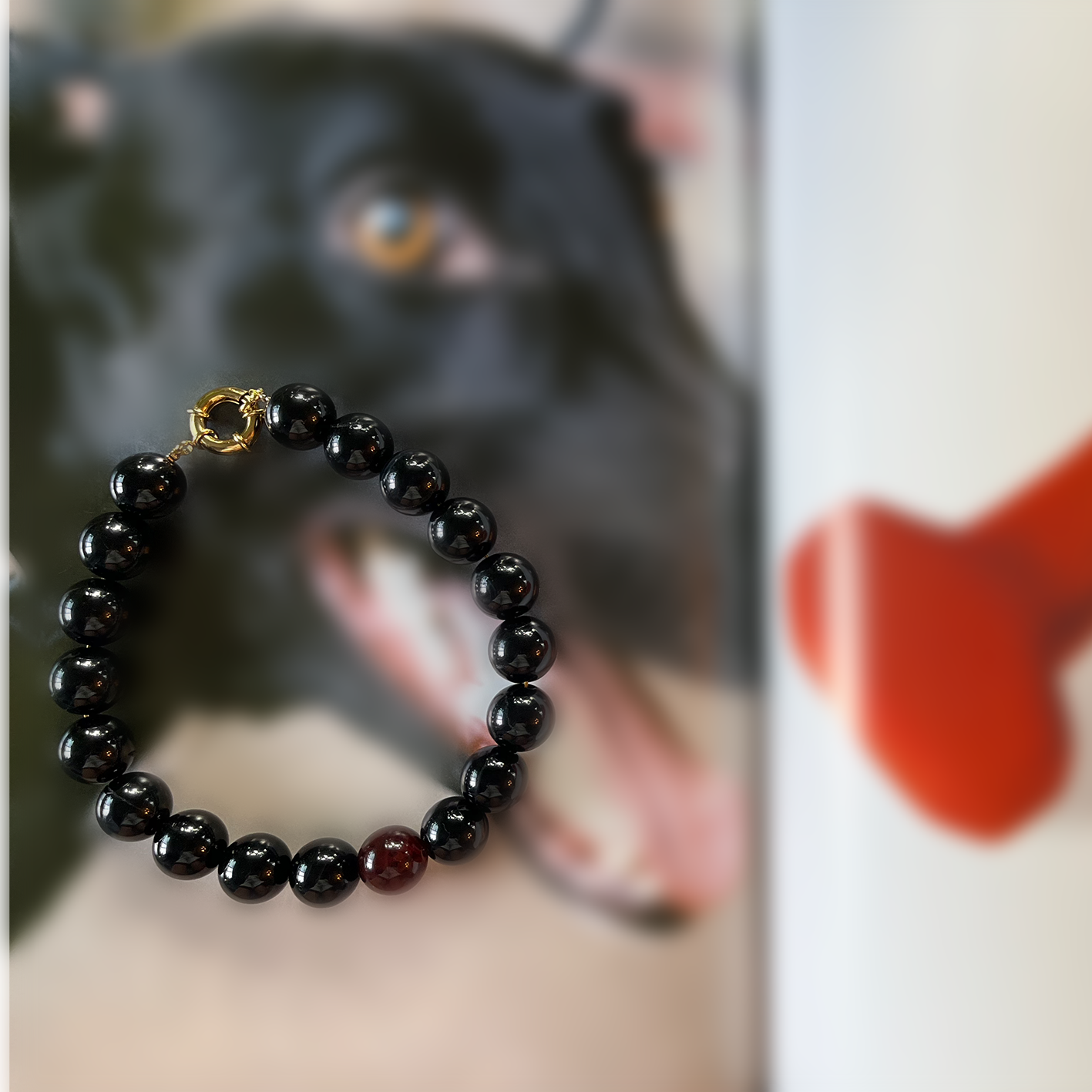 Pulsera de ónix negro con cuenta roja y broche dorado ROKA sobre fondo artístico desenfocado, joyería de piedras naturales